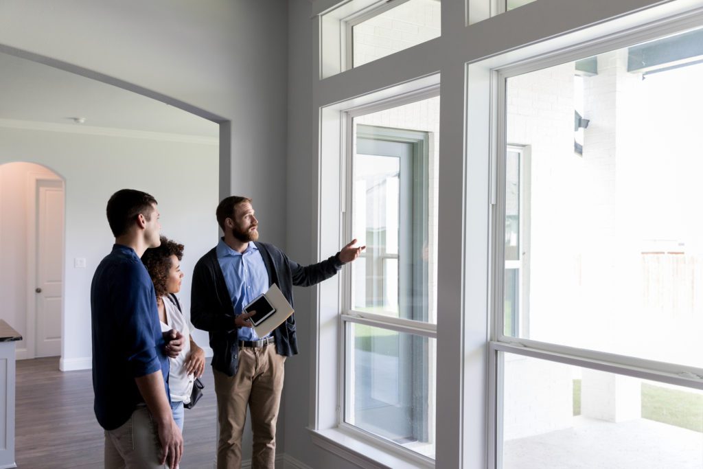 Confident male real estate agent gestures toward a beautiful view out the window of a new home. Interested potential homeowners attentively listen to the real estate agent.